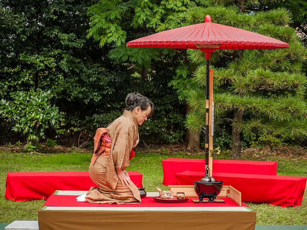 a picture of a lady in red and gold kimono under a paper umbrella, bowing to her tea utensils.